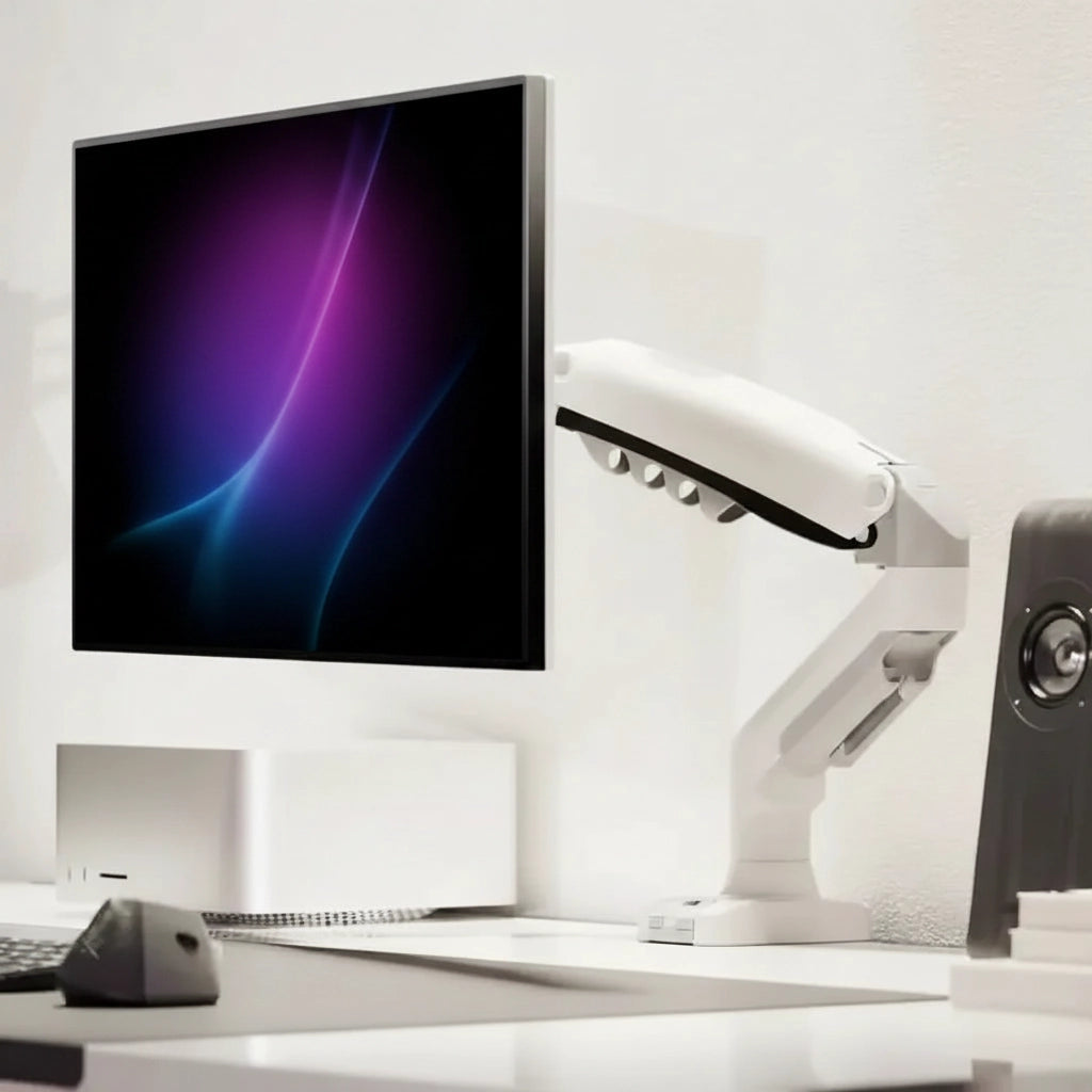 A white articulated desk monitor arm holding a black-framed computer monitor on a white desk, with a keyboard, mouse, and speakers in a bright minimalist office setting.